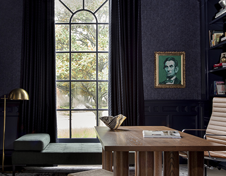 View of a custom wooden desk with a floor to ceiling iron arched window. The walls are a dark blue and there is a portait of Abraham Lincoln on the wall.